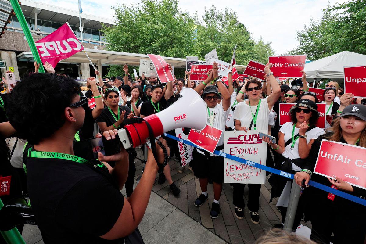 CUPE president Mark Hancock appears on a mobile phone screen as striking Air Canada flight attendants listen to a remote broadcast by megaphone by Hancock on the current situation, as they defy a back to work order at Vancouver International Airport in Richmond, British Columbia, Canada August 18, 2025. - REUTERSPIX