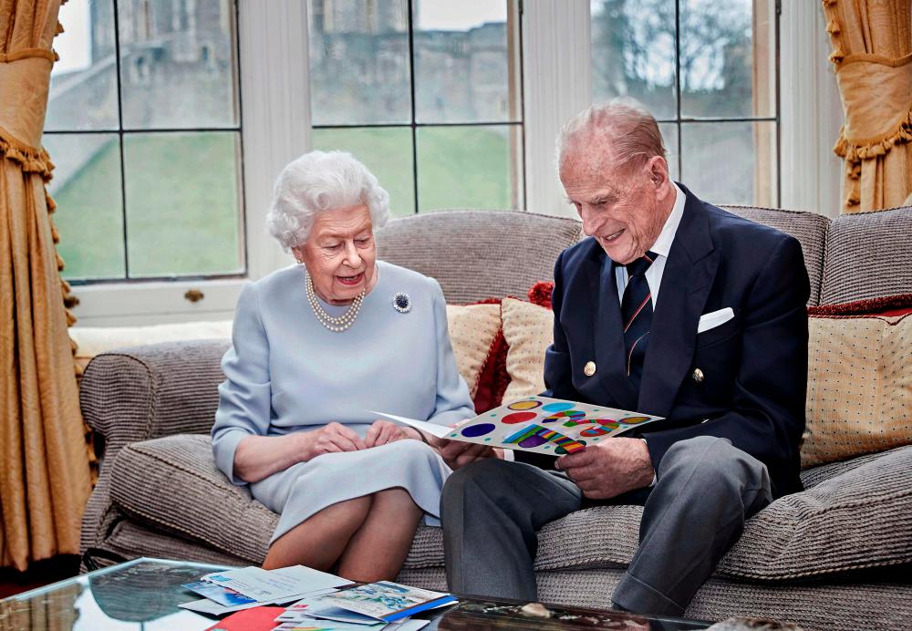 In this handout image released by Buckingham Palace on November 19, 2020, Britain’s Queen Elizabeth II (L) and Britain’s Prince Philip, Duke of Edinburgh (R) look at a homemade wedding anniversary card, given to them by their great grandchildren, Prince George, Princess Charlotte and Prince Louis in the Oak Room at Windsor Castle on November 17, 2020 in Windsor, west of London, ahead of their 73rd wedding anniversary on November 20. -AFP PHOTO / CHRIS JACKSON / GETTY IMAGES