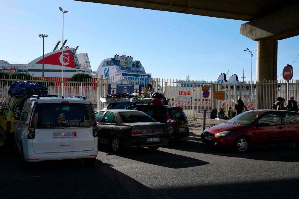 People sit near banners reading ‘Travellers and victims’ and ‘SOS’ in front of the entrance of Marseille-Fos Port, as they wait to take the ferry to Algeria, after they have been blocked for days since the strike against the government’s controversial pensions reform in Marseille, southeastern France, on March 16, 2023. AFPPIX