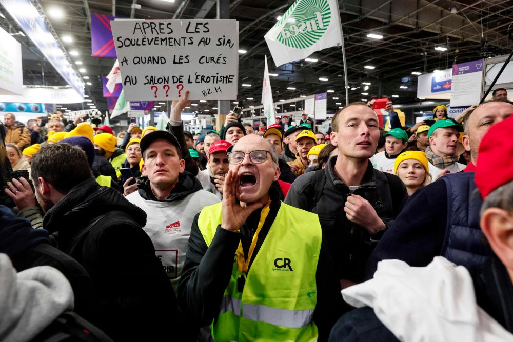A demonstrator shouts next to another holding a placard reading after the Soulevements at the SIA, when will we get priests at the Erotism salon? during a farmers' protest prior to the opening of the 60th International Agriculture Fair (Salon de l'Agriculture), at the Porte Versailles exhibition centre in Paris, on February 24, 2024. - AFPPIX
