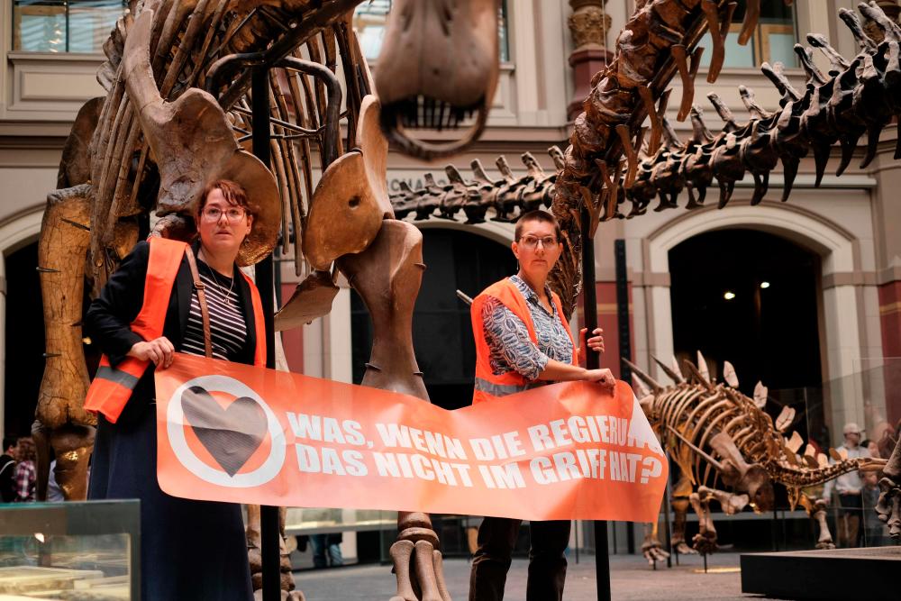 This handout picture released on October 30, 2022 by climate mouvement “Last Generation” shows two environmental activists of the group being glued to metal bars around a display of dinosaur skeletons and holding a banner reading “what if the government does not have it under control” in the Berlin’s Natural History Museum, in Berlin/AFPPix