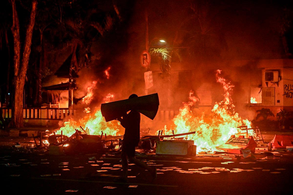 A protester carries looted goods on a street in front of a police headquarters that was set on fire and looted during demonstrations in Surabaya on August 31, 2025. Southeast Asia’s biggest economy has been rocked by protests in major cities, including the capital Jakarta, since April 29 after footage spread of a motorcycle taxi driver being run over by a police tactical vehicle during an earlier rally against low wages and financial perks for lawmakers. (Photo by Juni KRISWANTO / AFP)