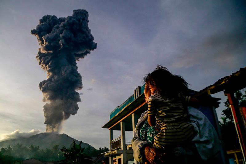 A woman and child look on at volcanic ash rising into the air during the eruption of Mount Ibu, as seen from Duono Village in West Halmahera, North Maluku province, on January 15, 2025. - AZZAM / AFP