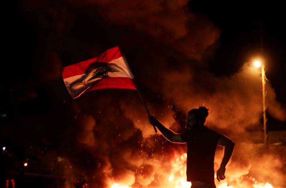 A Lebanese anti-government protester carries a Lebanese flag as he runs in front of burning tires blocking the main highway linking the city of Tripoli to Beirut at the coastal city of Byblos, north of the capital on Nov 13. — AFP