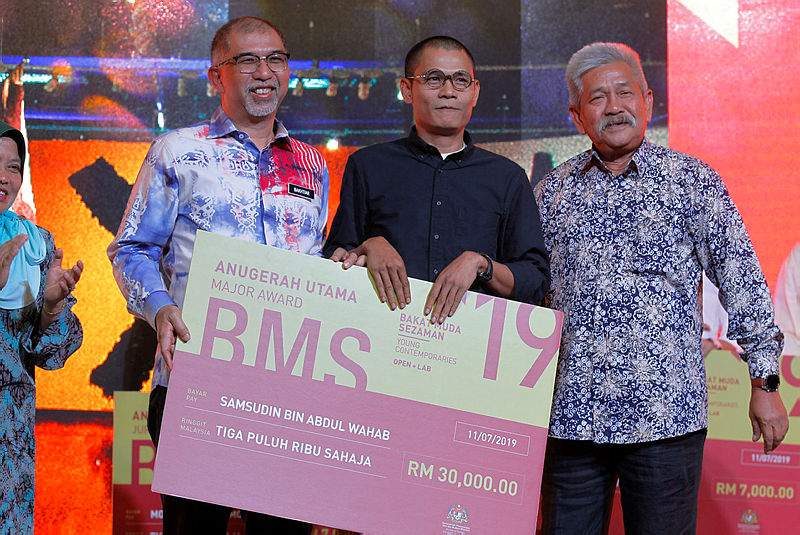 Deputy Minister of Tourism, Arts and Culture Muhammad Bakhtiar Wan Chik (L) presents a mock cheque to the recipient of the Young Contemporary Award (BMS19), Samsudin Abdul Wahab (C), on July 11, 2019. — Bernama