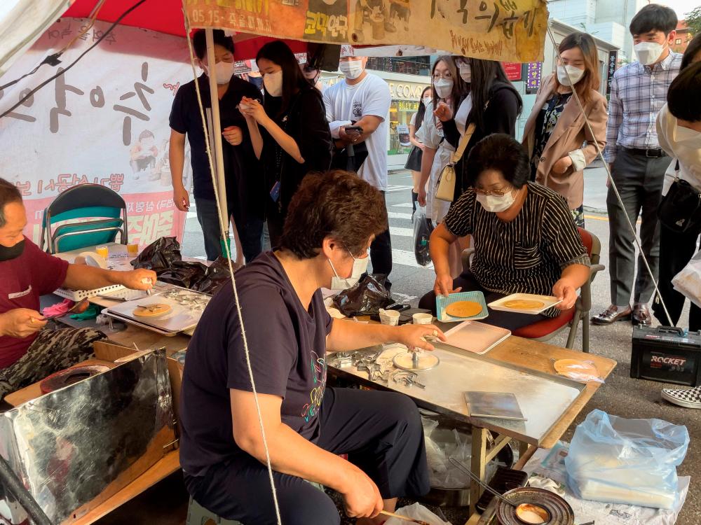 In this picture taken in Seoul on October 10, 2021, customers wait in line as street vendor Jung Jung-soon (C) and her husband Lim Chang-joo (L) sell freshly made dalgonas, a crisp sugar candy featured in the Netflix smash hit series Squid Game, for which they were hired to be on set to make during production. AFPpix