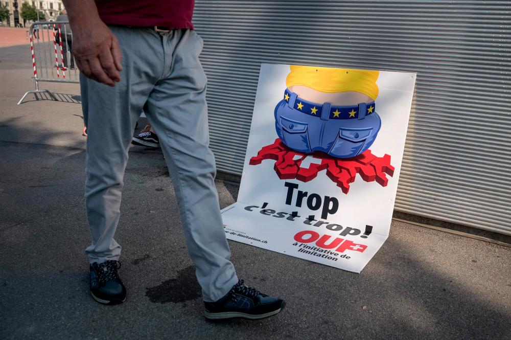 A picture taken on Sept 13, 2020 during a Sunday market in Geneva shows an electoral poster by right-wing Swiss People’s Party (SVP) showing a cartoon worker wearing a belt studded with EU stars, crushing the red and white map of Switzerland with his wide rear end that translates from French as “Too much is too much!” to advertise their nationwide initiative demanding that the government scrap the freedom of movement agreement with the EU within a year. — AFP