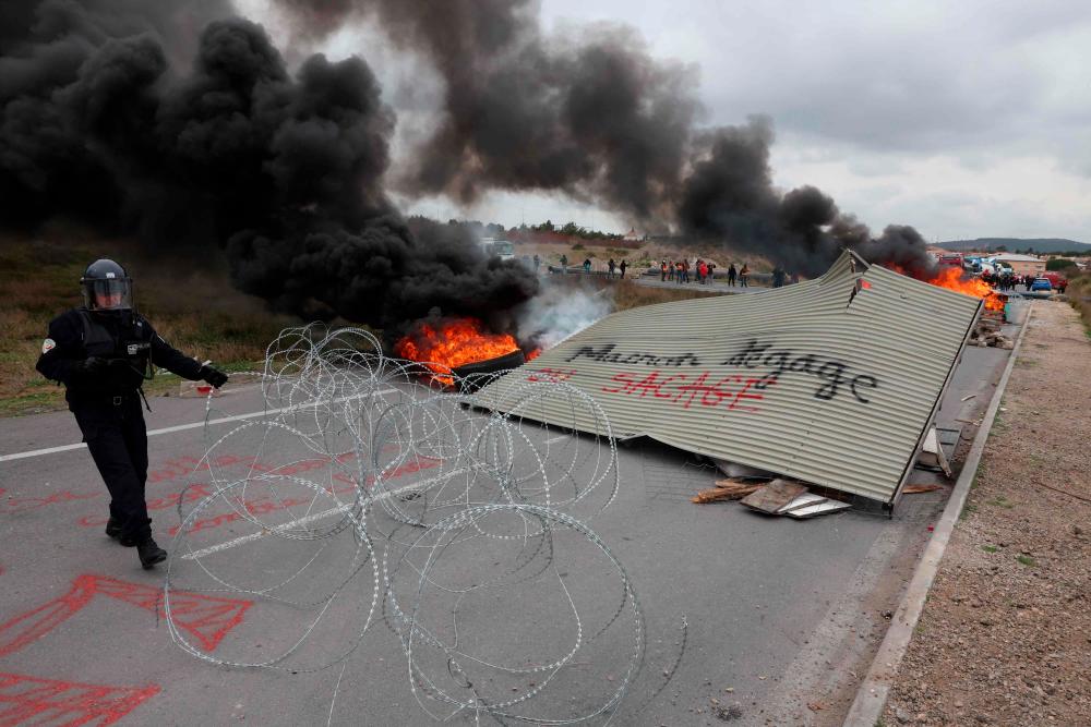 A police officer in riot gear removes barbed wire next to a panel reading “Macron, get out or else ransacking”, at a barricade and fires as protesters block the access to the Frontignan oil depot a week after the government pushed a pensions reform through parliament without a vote, using the article 49.3 of the constitution, in Frontignan, southern France, on March 24, 2023. AFPPIX