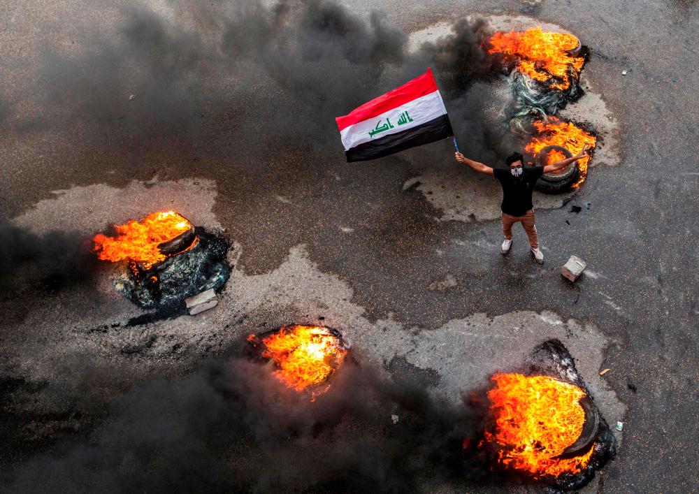 An Iraqi protester wearing a face bandana and waving a national flag poses past burning tires blocking a road during a demonstration in the southern city of Basra on Nov 17, 2019, as protesters cut-off roads and activists call for a general strike. — AFP
