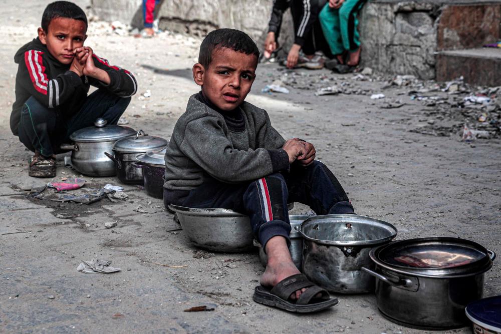 Boys sit with empty pots as displaced Palestinians queue for meals provided by a charity organisation ahead of the fast-breaking iftar meal during the Muslim holy month of Ramadan, in Rafah in the southern Gaza Strip on March 16, 2024 - AFPPIX