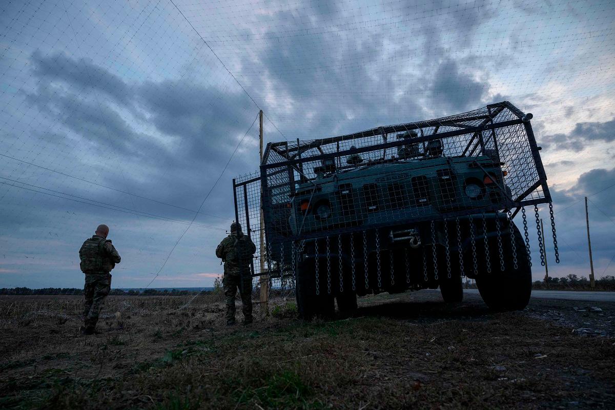 Ukrainian soldiers stand before an armoured military vehicle surrounded by a cage to protect against drone attacks on a road near Oleksandrivka, on October 6, 2025 amid the Russian invasion of Ukraine. - Ed JONES / AFP