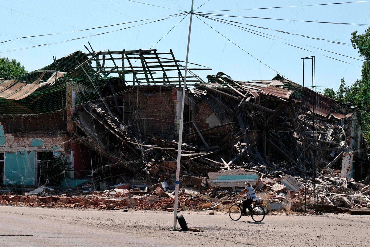 In this handout photograph taken and released by the press service of the 65th Mechanized Brigade of Ukrainian Armed Forces on July 15, 2025, a woman rides a bicycle near damaged buildings and anti-drone nets in Orikhiv, Zaporizhzhia region, amid the Russian invasion of Ukraine. - AFPPIX/65th Mechanized Brigade of Ukrainian Armed Forces