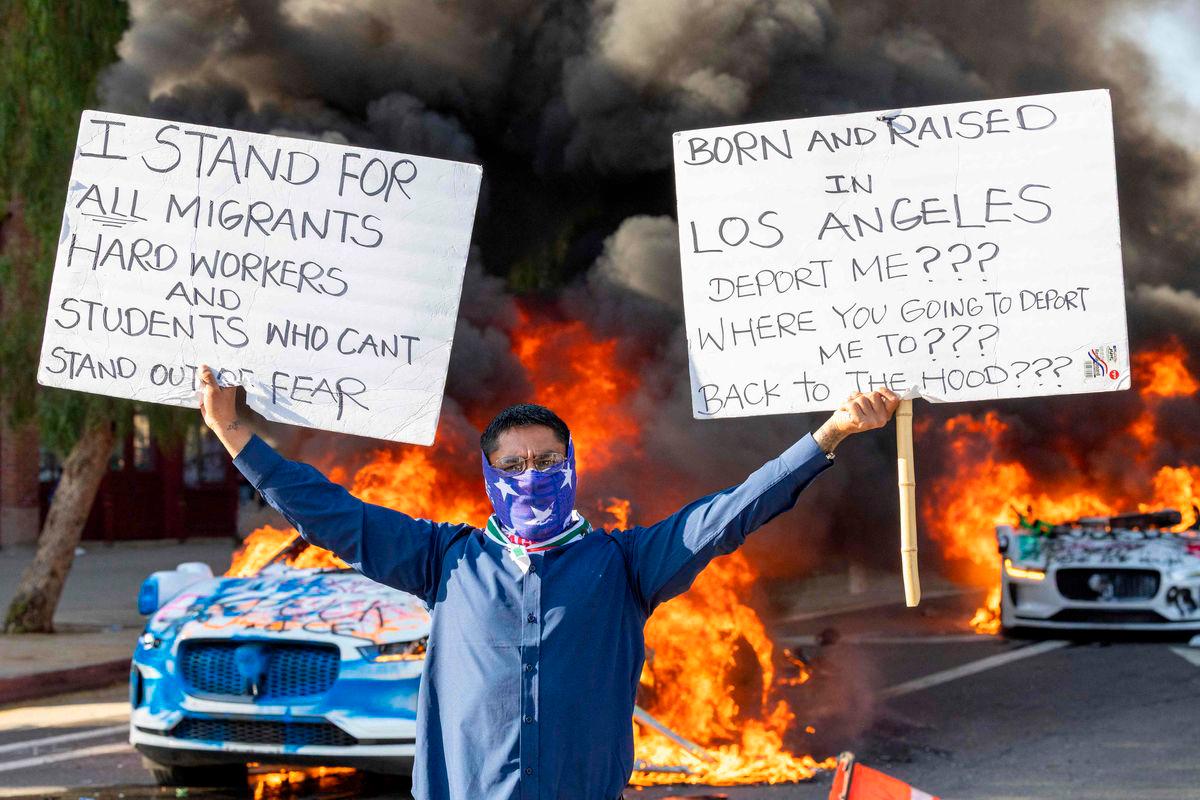 A man holds signs as a Waymo vehicles burn, as protesters clash with law enforcement in the streets surrounding the federal building during a protest following federal immigration operations in Los Angeles, California, on June 8, 2025.